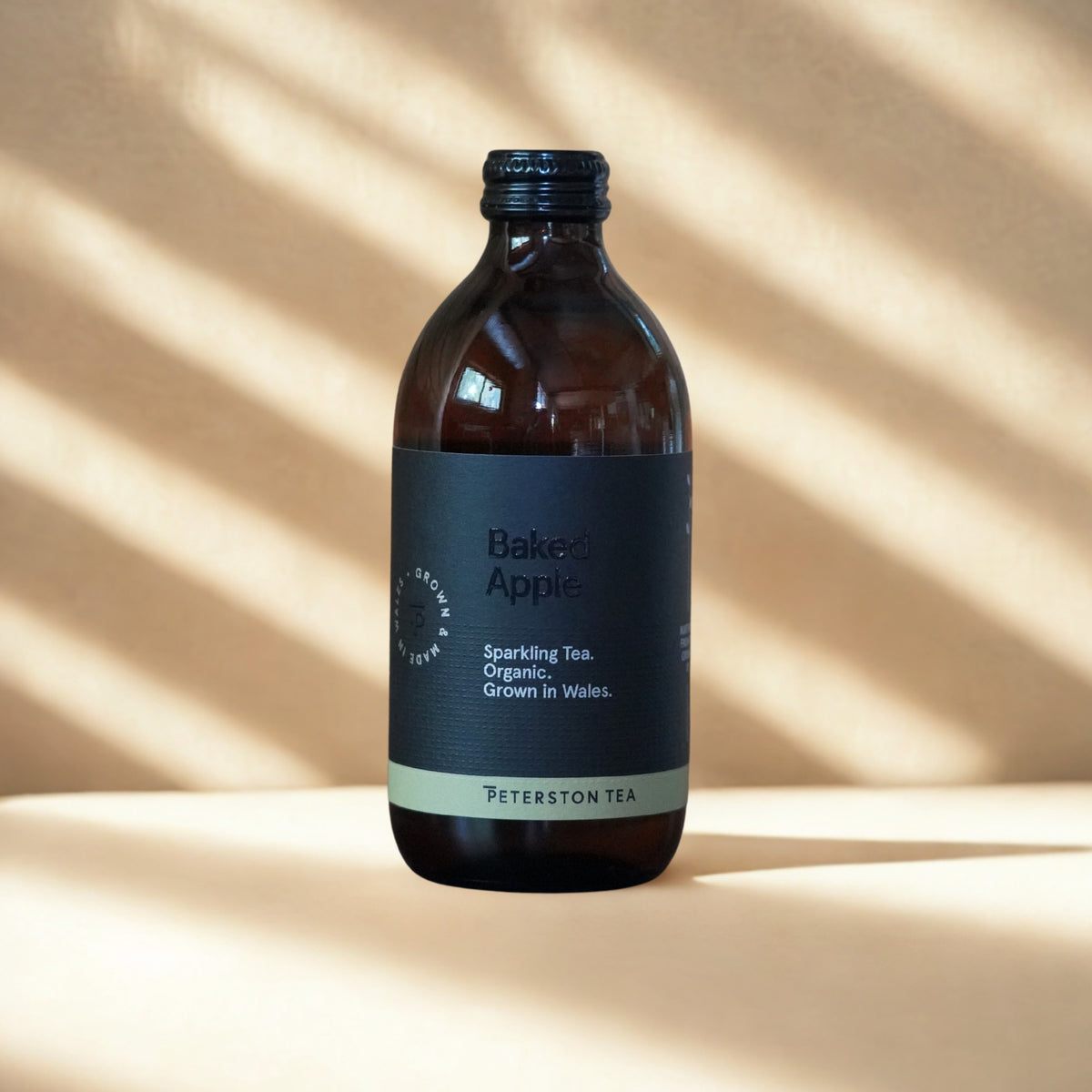 A bottle of Peterston Tea Baked Apple Sparkling Tea, made from organic tea grown in Wales, shown on a beige surface with diagonal shadow patterns. The bottle is dark amber glass with a black cap and a dark charcoal gray label featuring a light green band at the bottom.