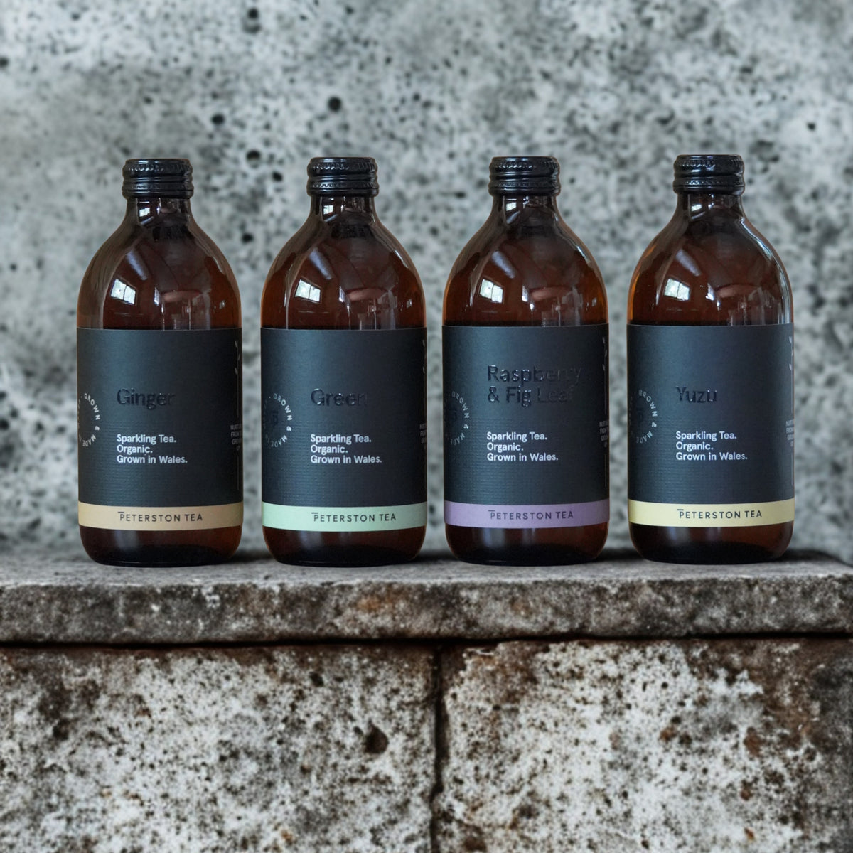 Four amber glass bottles of Peterston Tea sparkling tea lined up on a stone ledge against a textured concrete wall. The bottles have black labels with different colored accents and are labeled Ginger, Green, Raspberry & Fig, and Yuzu.