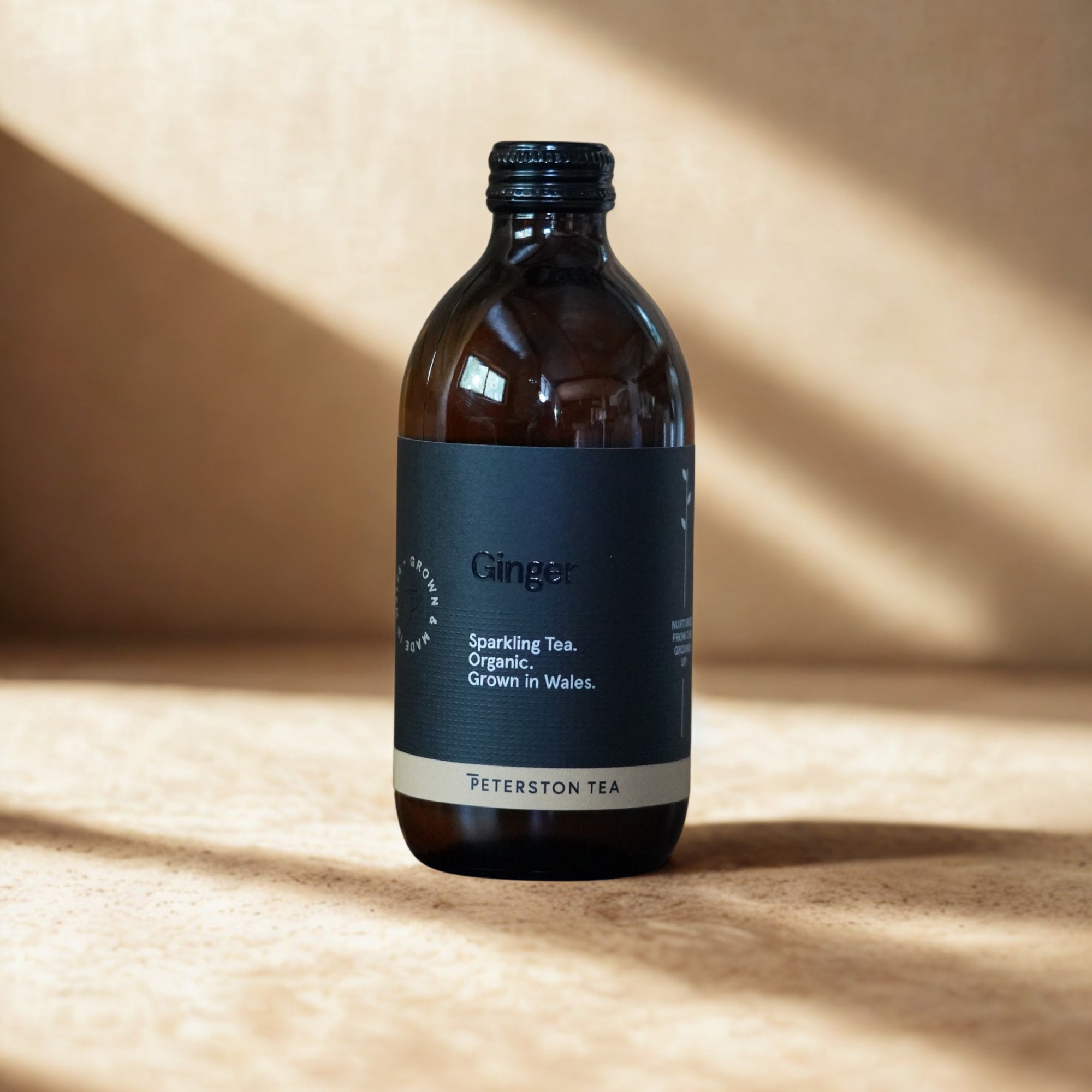 A bottle of Peterston Tea Ginger Sparkling Tea, organic, grown in Wales, shown on a textured beige surface with dramatic diagonal shadows.