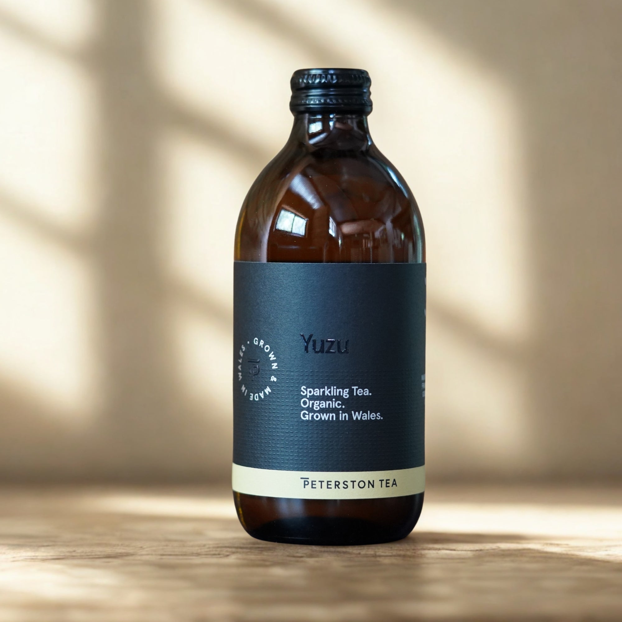 A bottle of Peterston Tea Yuzu sparkling tea, made from an amber glass bottle with a dark navy blue label and a cream band at the bottom, set on a wooden surface with soft window shadow lighting.